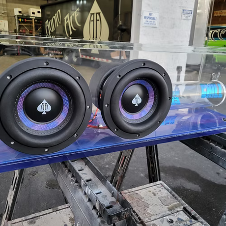 Two black Audio Ace subwoofer speakers with a logo on a blue surface, with a store interior in the background.